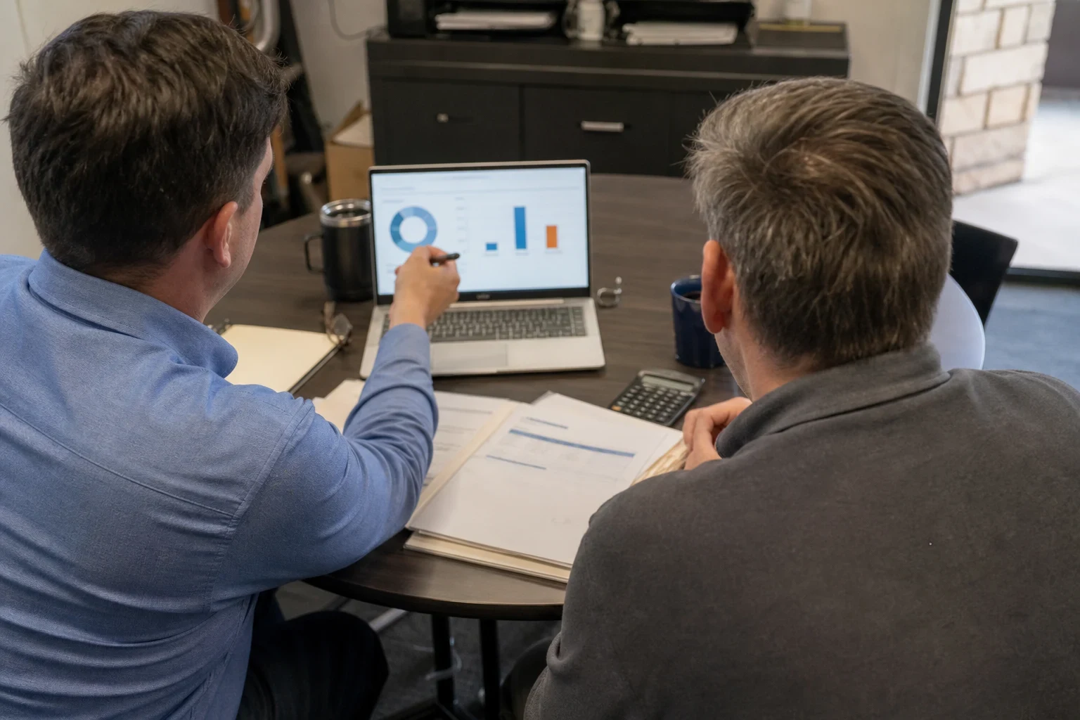 Agent in a blue button-down points a pen at charts on a laptop while a client in a gray sweater looks on at a round office table with papers and a calculator