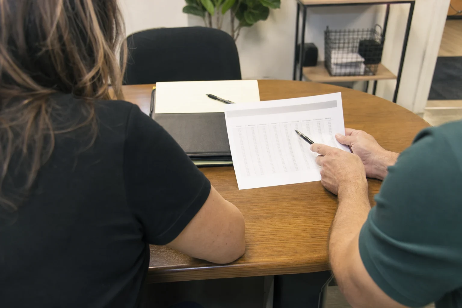 Two people seated at a round wooden table reviewing a printed page of figures, one pointing with a pen, beside a black portfolio and notepad in a small office.