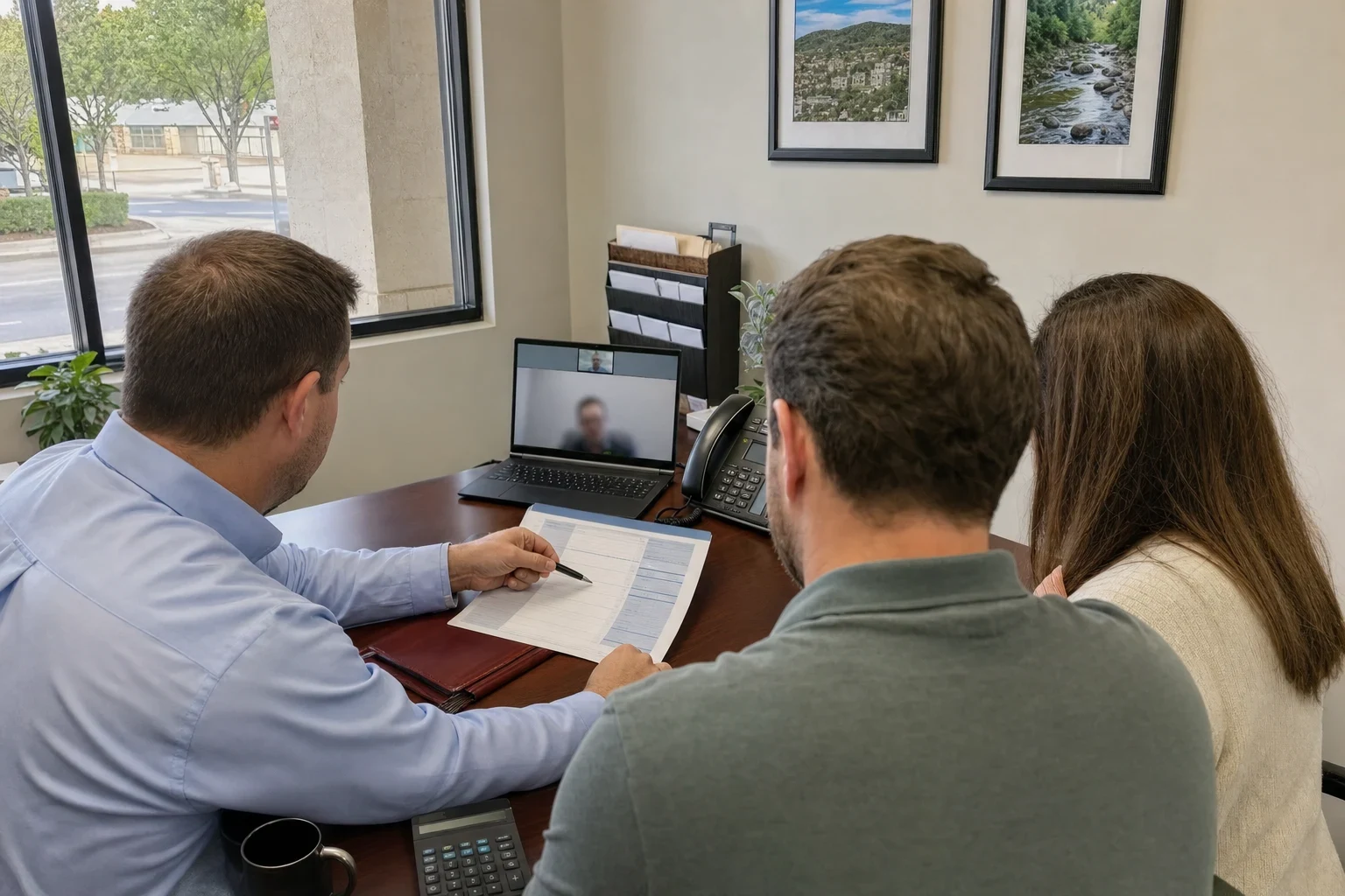 Insurance agent in a light blue shirt points a pen at a printed quote sheet while a couple sits beside him at an office desk with a laptop, phone and window